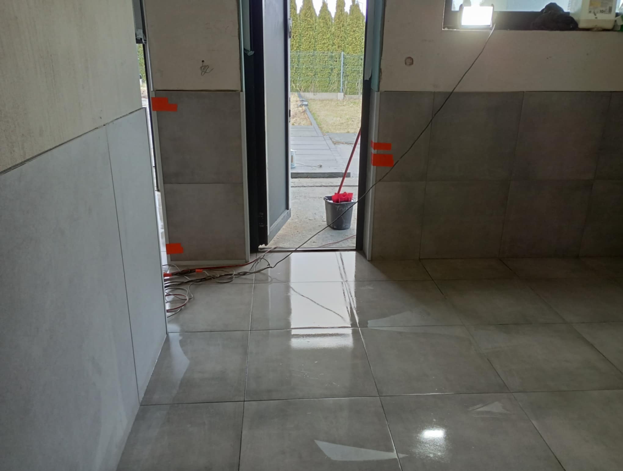 Świeżo ułożone, błyszczące, szare płytki podłogowe w pomieszczeniu z otwartym przejściem na zewnątrz. Widoczne pomarańczowe kliny dystansowe i wiadro z narzędziami.