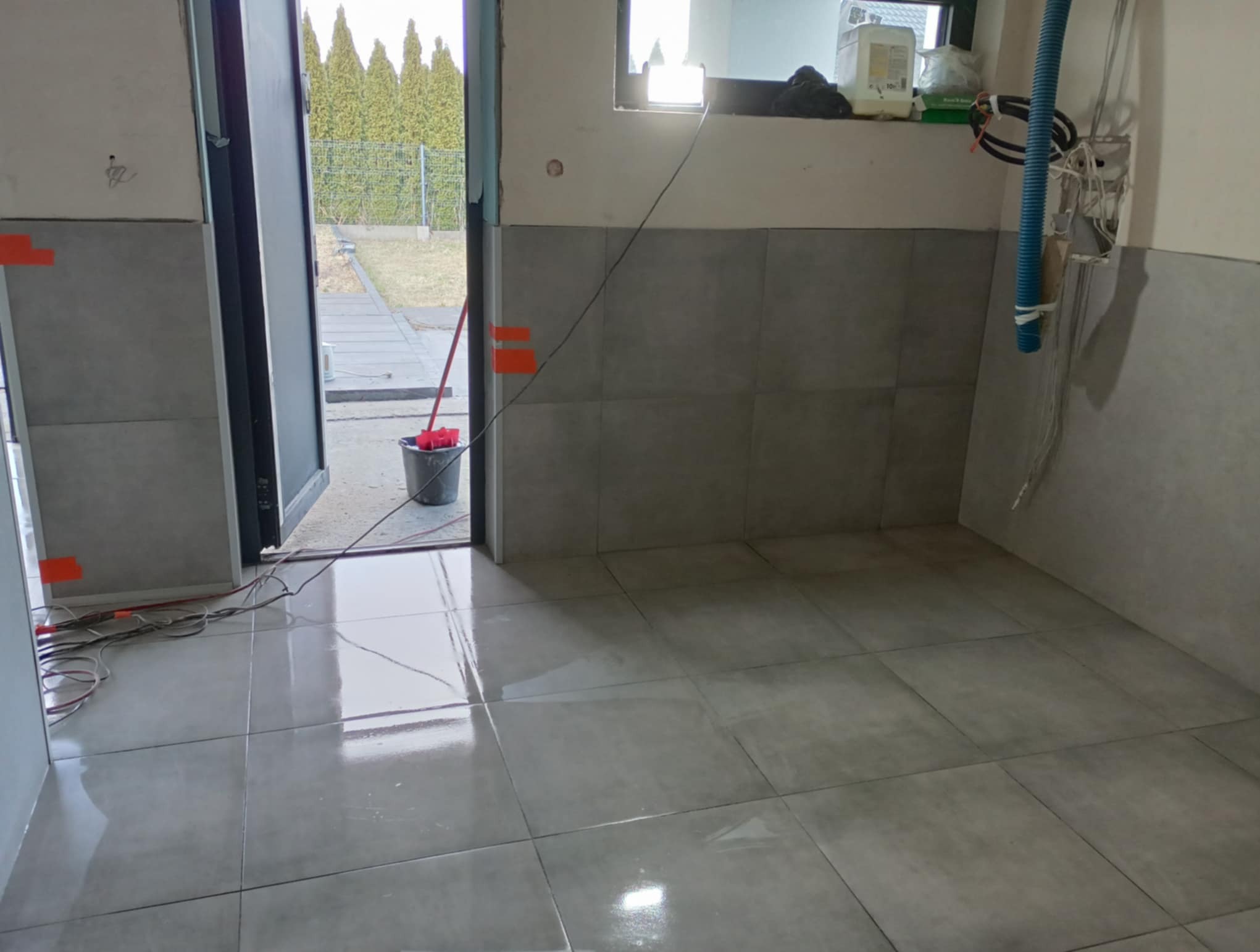 Ułożone szare płytki podłogowe i częściowo ścienne w pomieszczeniu w trakcie prac wykończeniowych. Widoczne narzędzia i materiały budowlane.