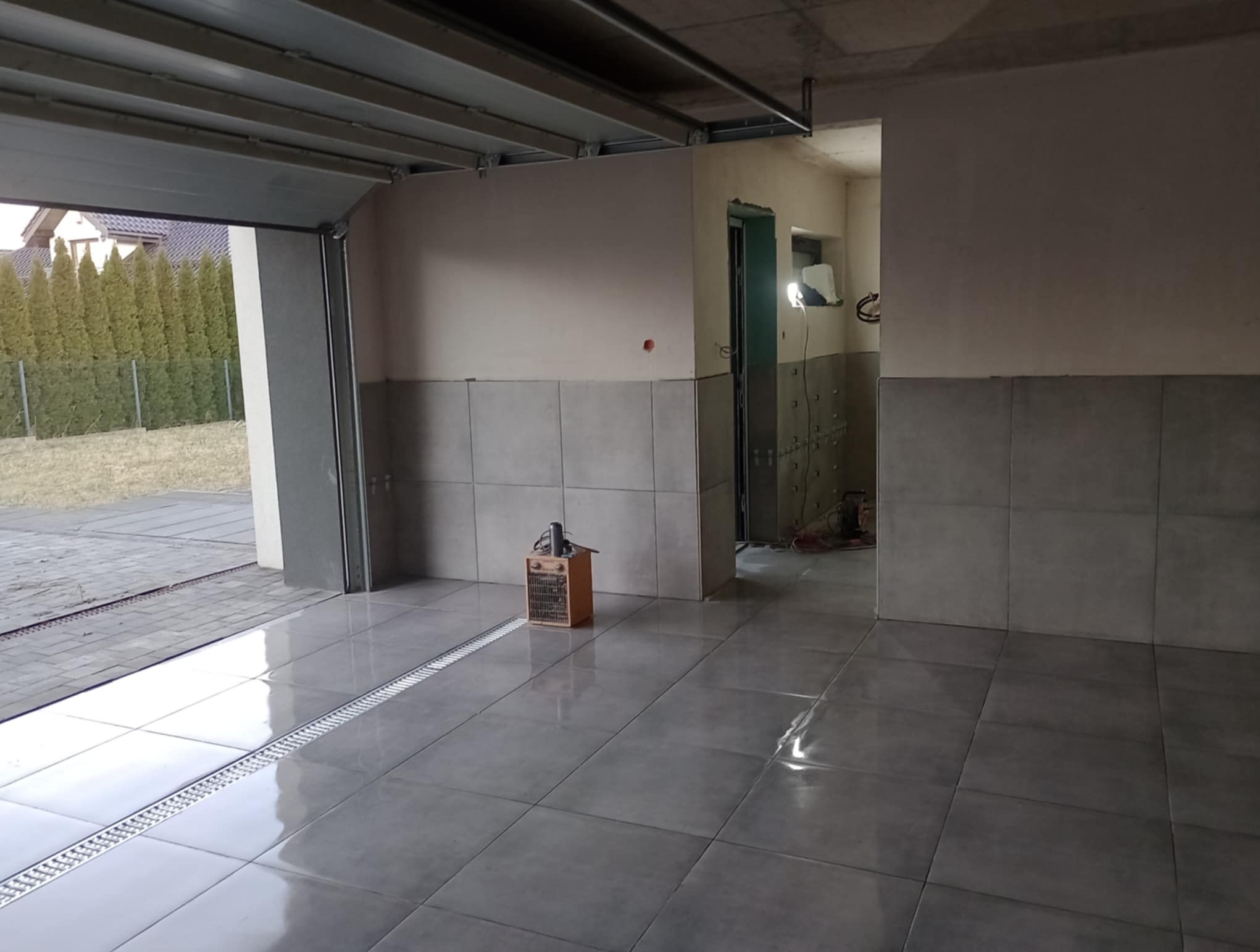 Wykończony garaż z szarymi płytkami na podłodze i ścianach do połowy wysokości. Widoczny odpływ liniowy i grzejnik elektryczny. Otwarta brama garażowa.