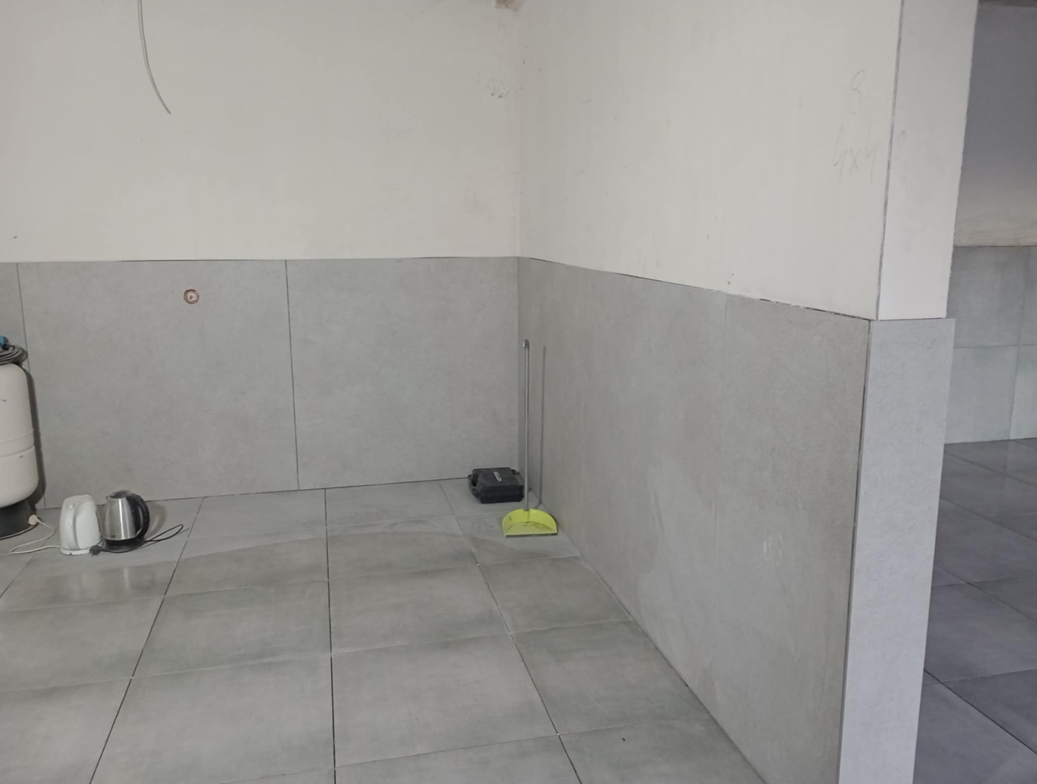 Szare płytki na ścianach i podłodze w rogu pomieszczenia. Widoczny sprzęt AGD i narzędzia. Minimalistyczne wykończenie wnętrza w odcieniach szarości.