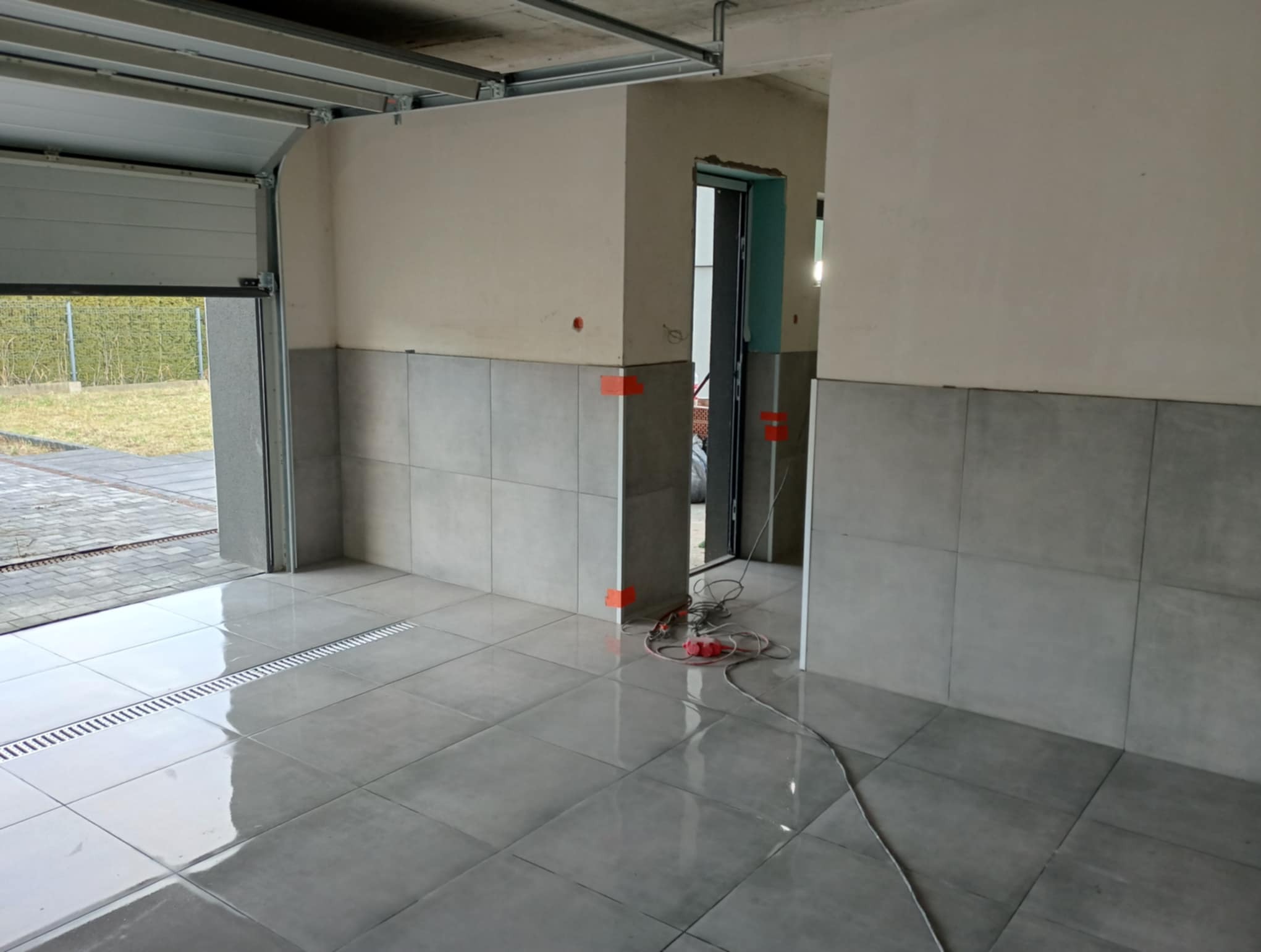 Wykończone płytkami podłoga i ściany w garażu z otwartą bramą. Widoczne narzędzia i kable. Szare płytki, jasne ściany. Etap prac wykończeniowych.