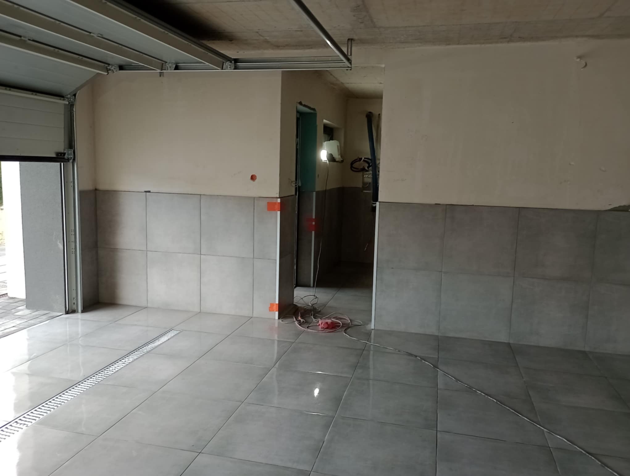 Wykończony garaż z szarymi płytkami na ścianach do połowy wysokości i na podłodze z odpływem liniowym. Widoczne niedoskonałości na ścianach i surowy betonowy sufit.