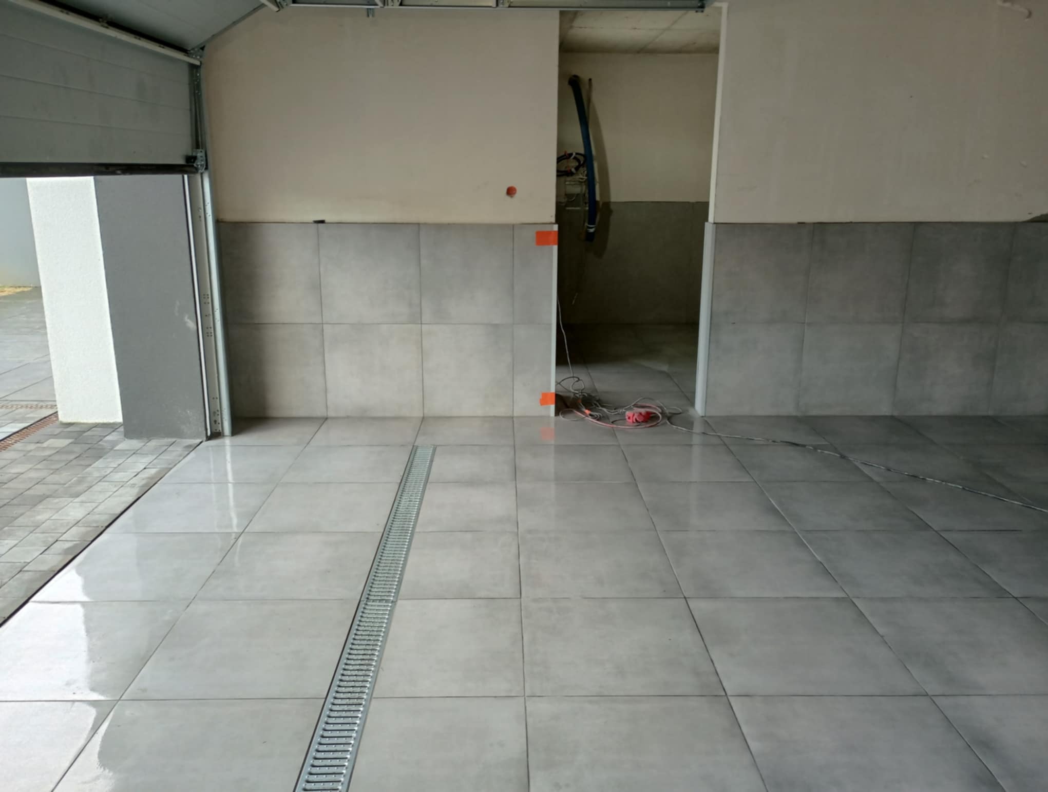 Wykończona podłoga z szarych płytek w garażu z odpływem liniowym. Częściowo wykończone ściany z szarymi płytkami. Widoczne kable i otwarty wjazd do garażu.
