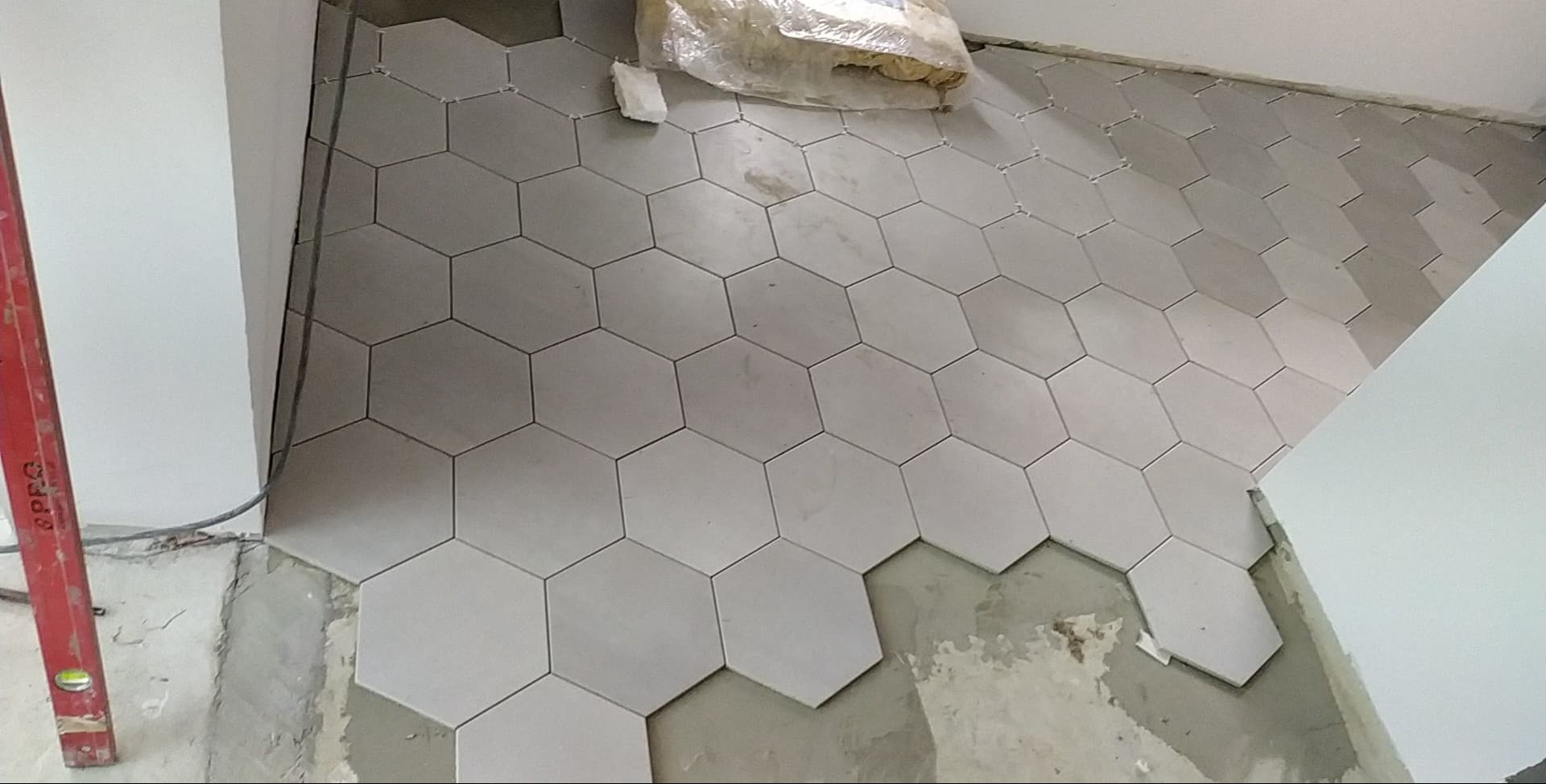 Układanie jasnych, heksagonalnych płytek podłogowych w trakcie prac wykończeniowych, widoczna poziomica i worek z zaprawą.
