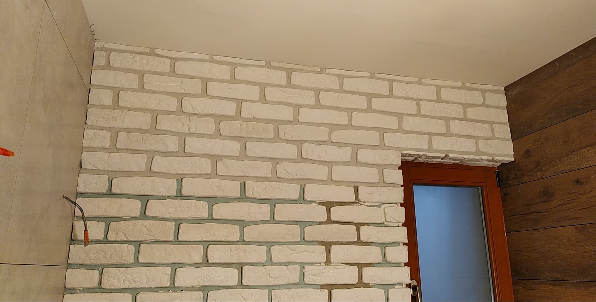 Ściana z białej cegły dekoracyjnej, fragment okna z brązową ramą, oraz fragment ściany wyłożonej drewnem. Widoczne przewody elektryczne.