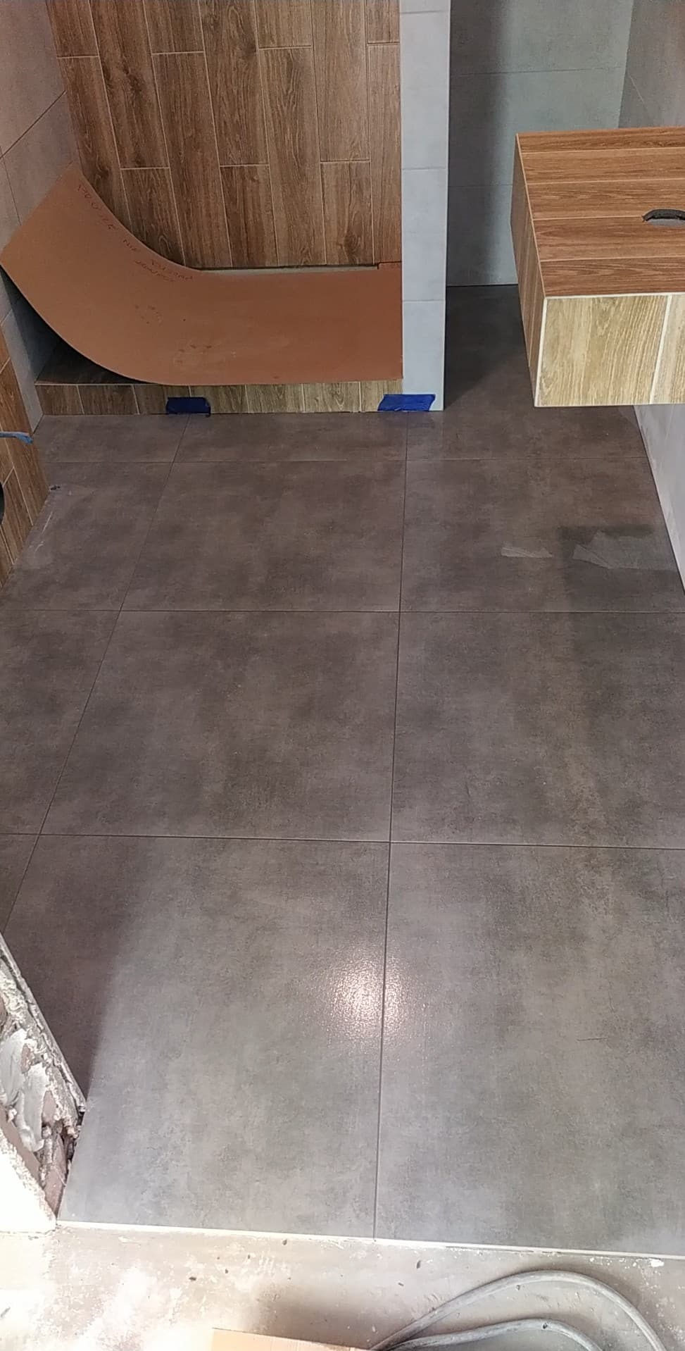 Nowoczesna łazienka po remoncie: szare płytki podłogowe, drewnopodobne płytki na ścianie prysznica i szafka pod umywalkę w podobnym stylu. Widok z góry.