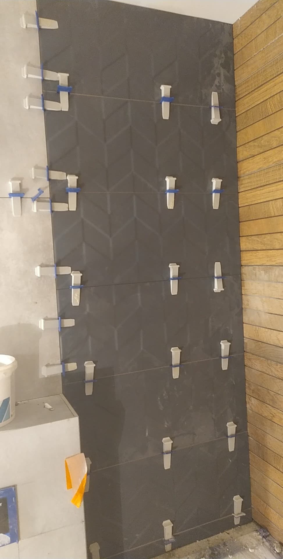 Układanie ciemnoszarych płytek z wzorem w jodełkę na ścianie, z użyciem białych klipsów poziomujących i sznurka. Fragment drewnianej ściany w tle.