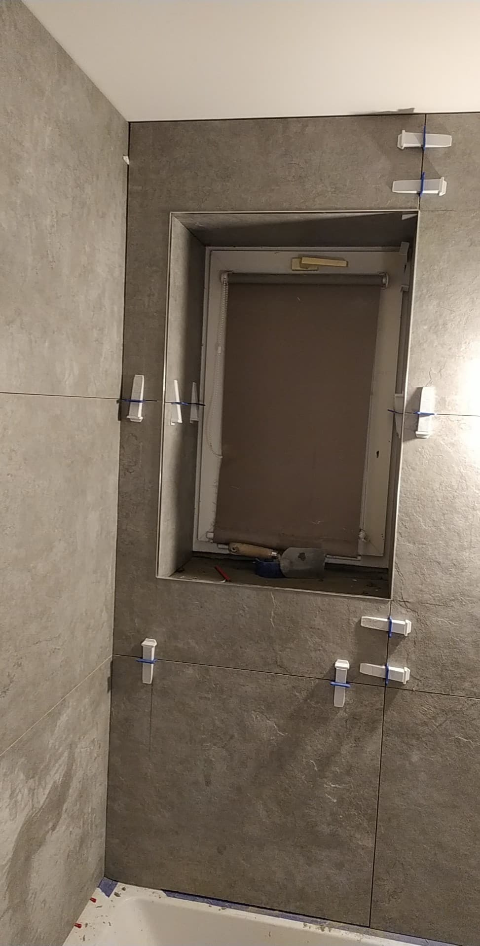 Szare płytki ścienne w trakcie montażu, z krzyżykami dystansowymi i narzędziami widocznymi w niszy okiennej. Ujęcie z bliska, prezentujące etap prac wykończeniowych.