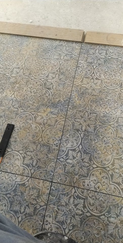 Ułożone płytki podłogowe z dekoracyjnym wzorem, widoczne fugi i listwy przypodłogowe, narzędzie glazurnicze w rogu kadru.