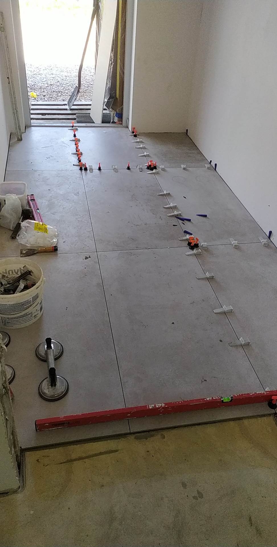 Szare płytki podłogowe w trakcie układania, z użyciem poziomicy i klipsów dystansowych, w jasnym pomieszczeniu z widocznym wejściem.