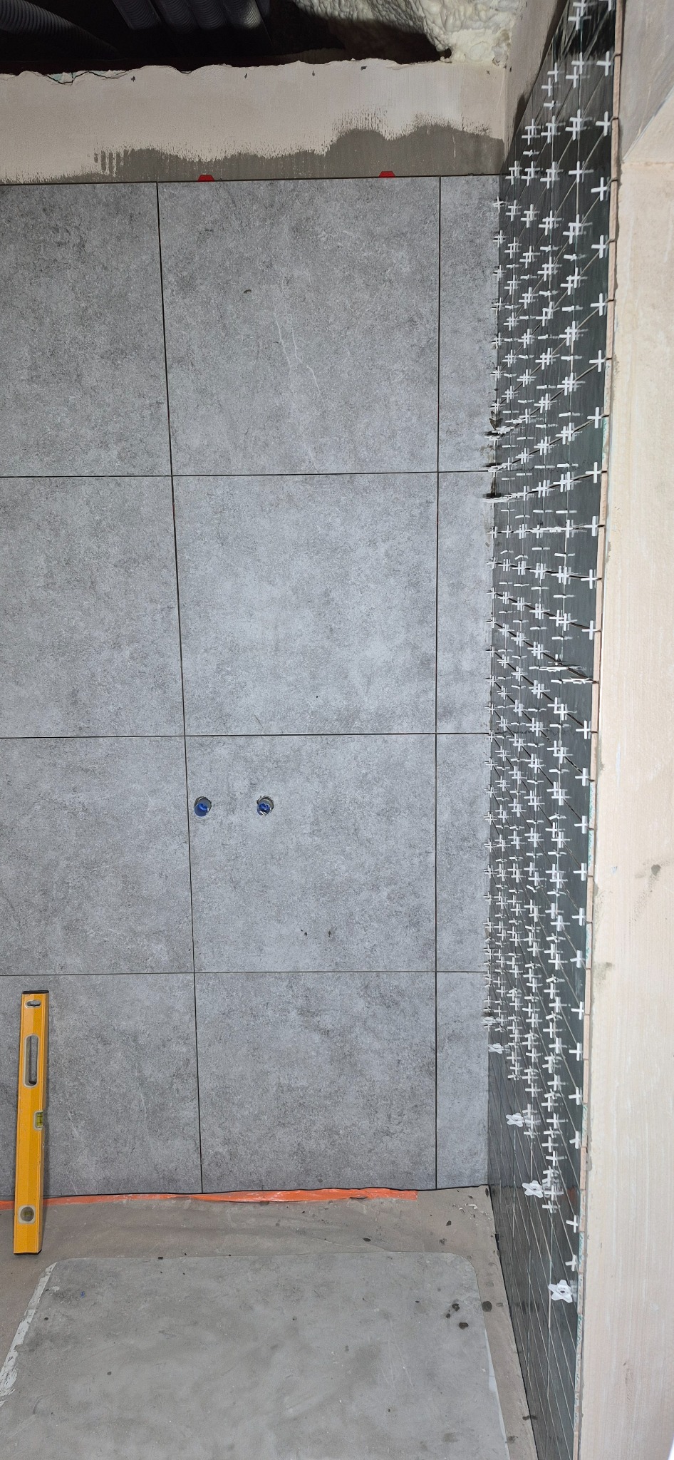 Ściana z szarych, kwadratowych płytek, częściowo ułożonych, z krzyżykami dystansowymi. Poziomica przy ścianie. Widoczne instalacje. Surowy stan wykończenia.