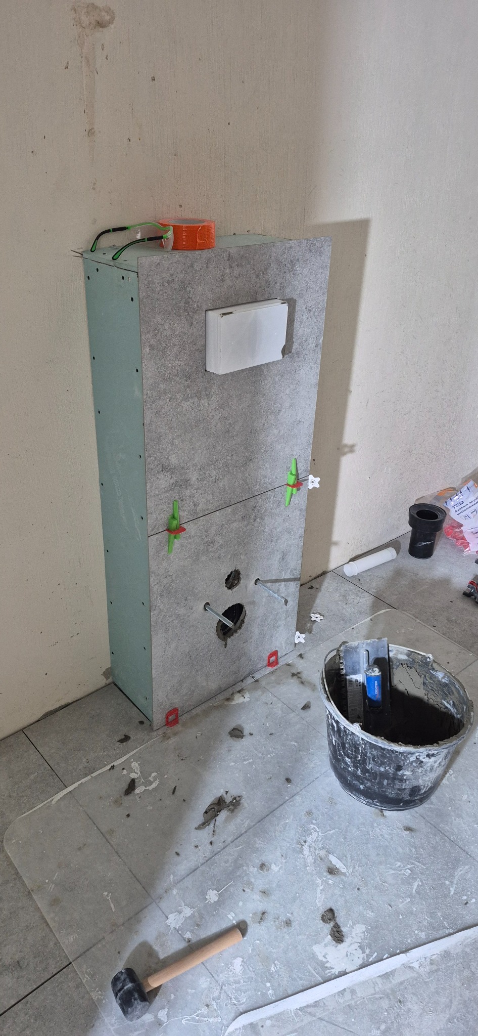 Montaż stelaża podtynkowego WC z płytkami imitującymi kamień. Widoczne narzędzia i akcesoria glazurnicze, poziomowanie płytek. Prace wykończeniowe w łazience.