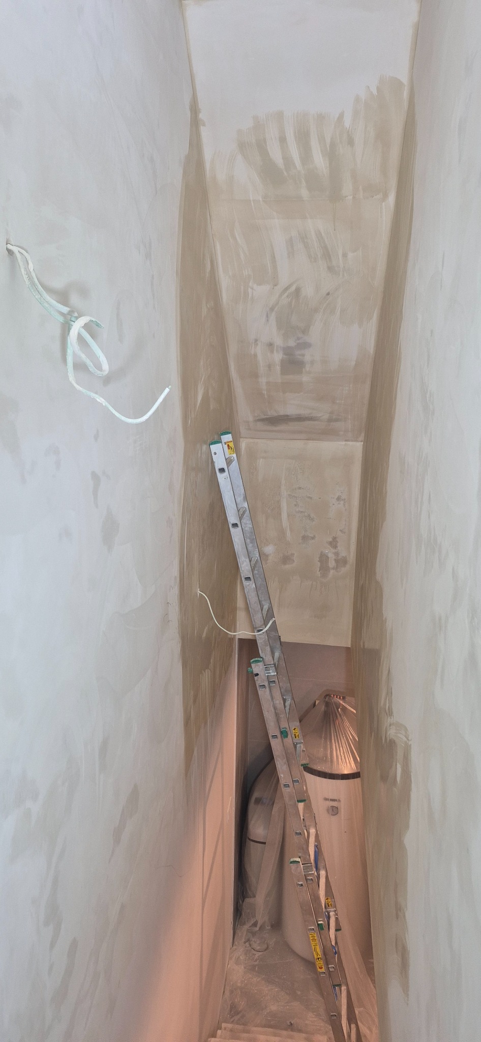 Ściana w trakcie szpachlowania z widocznymi przewodami elektrycznymi i drabiną opartą o ścianę. Widoczne ślady po szpachli na ścianie i suficie.