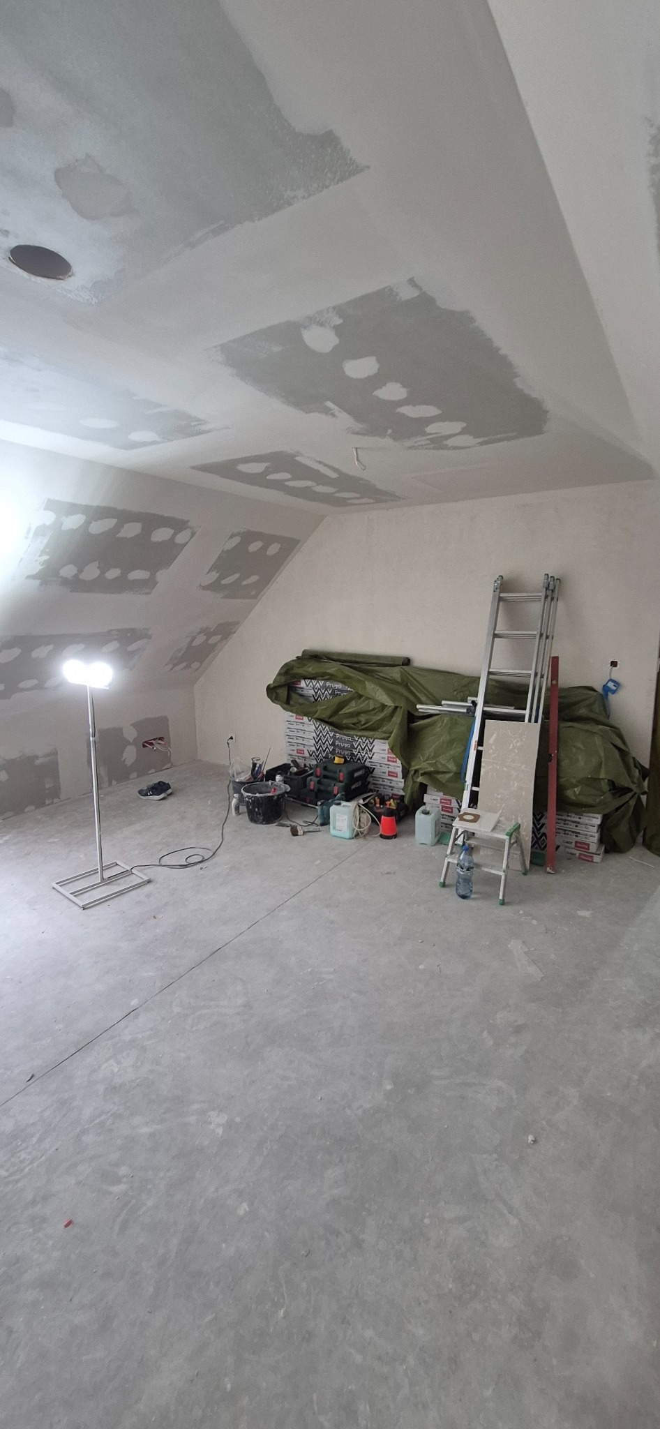 Poddasze w trakcie prac wykończeniowych: szpachlowana sufit i ściany, narzędzia, drabina, oświetlenie robocze. Widok na poddasze w trakcie szpachlowania.