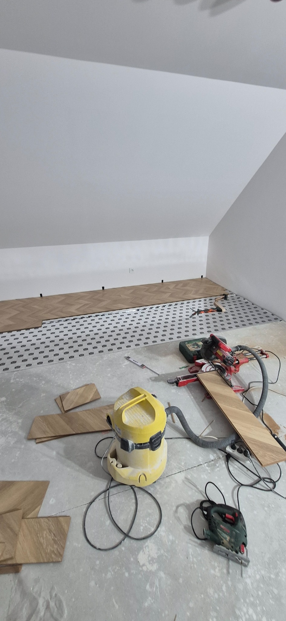 Układanie parkietu w jodełkę w pokoju na podkładzie. Widoczny odkurzacz przemysłowy, piła do cięcia paneli i wyrzynarka. Częściowo ułożona podłoga.