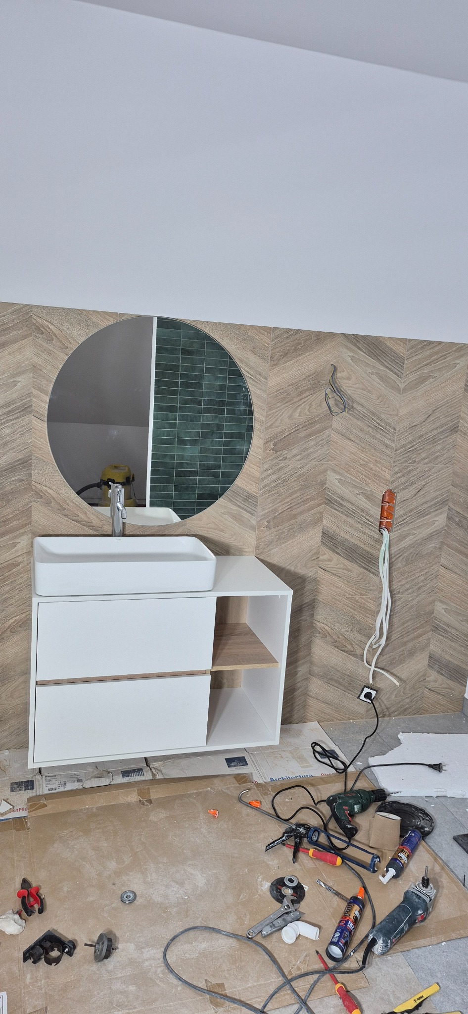 Łazienka w trakcie wykończenia: biała szafka z umywalką, okrągłe lustro, płytki w jodełkę na ścianie, widoczne narzędzia i kable. Etap prac wykończeniowych.