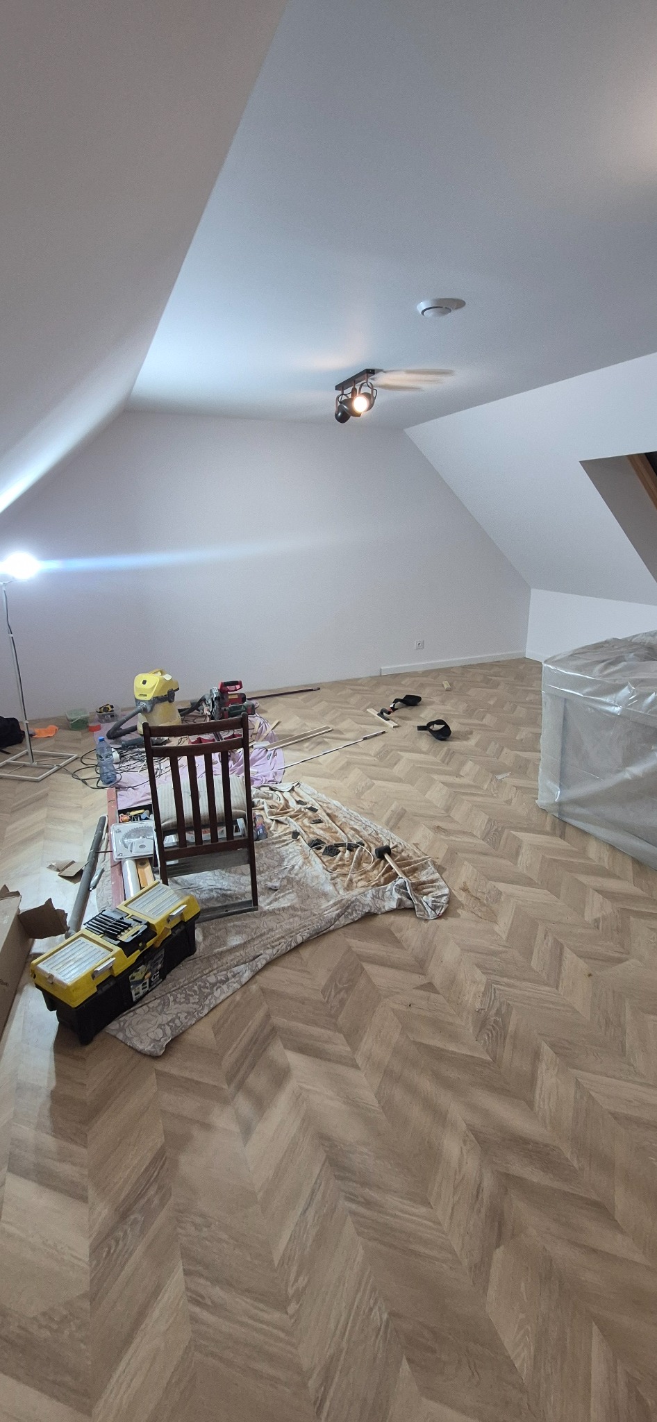 Ukończona podłoga w jodełkę w pokoju na poddaszu, narzędzia pracy i krzesło w tle. Widoczne skosy sufitu i lampa sufitowa.