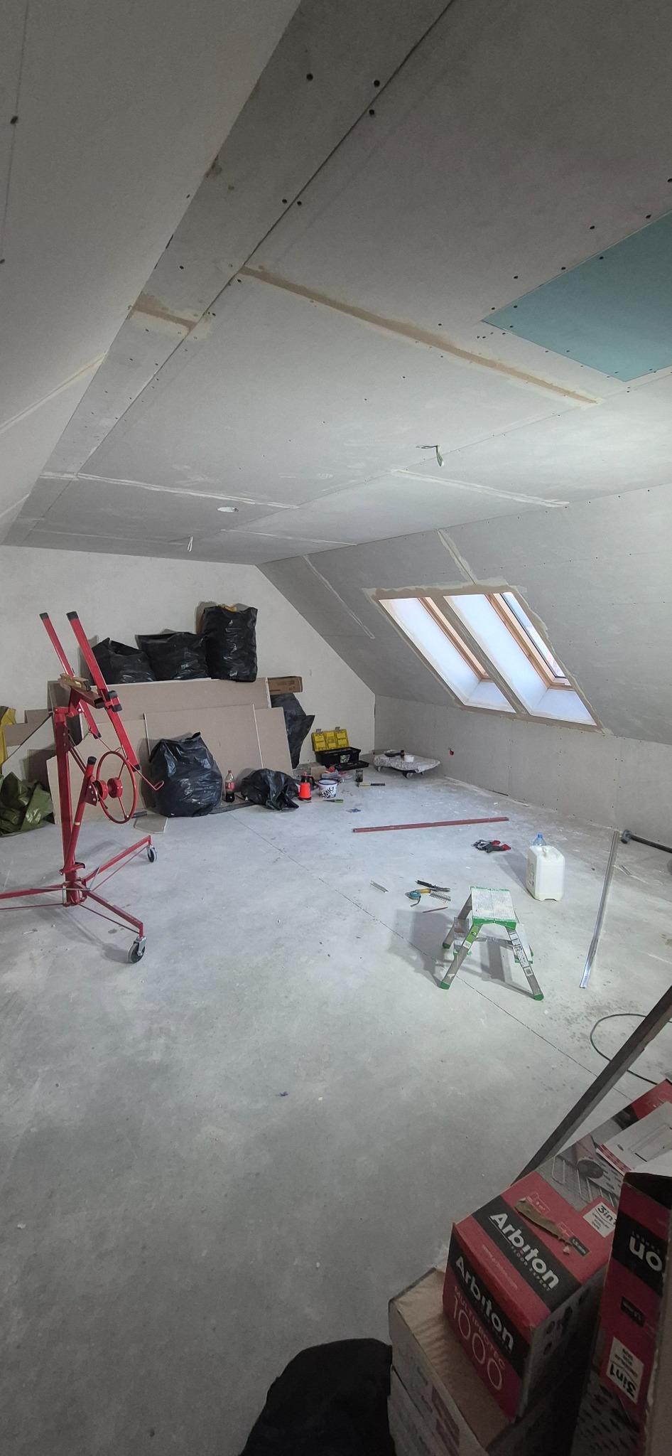 Poddasze w trakcie prac wykończeniowych: zamontowane płyty gipsowo-kartonowe na skosach, widoczne okna dachowe, narzędzia i materiały budowlane. W tle czarne worki na śmieci.