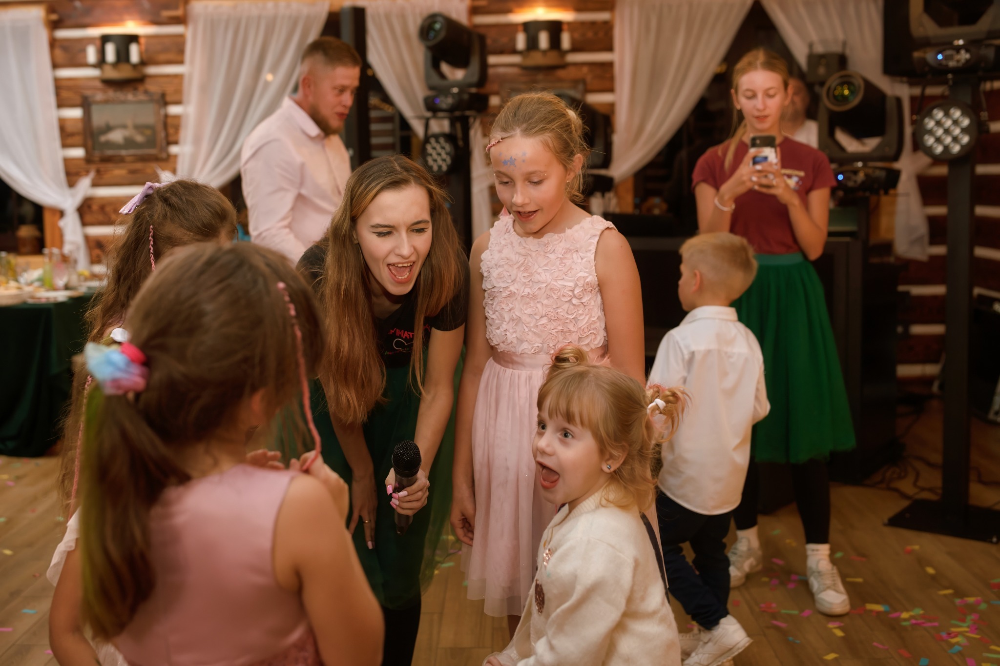 Animator z mikrofonem prowadzi zabawę dla grupy dzieci w sali z drewnianymi elementami dekoracyjnymi i oświetleniem eventowym. Dziewczynki ubrane w sukienki.