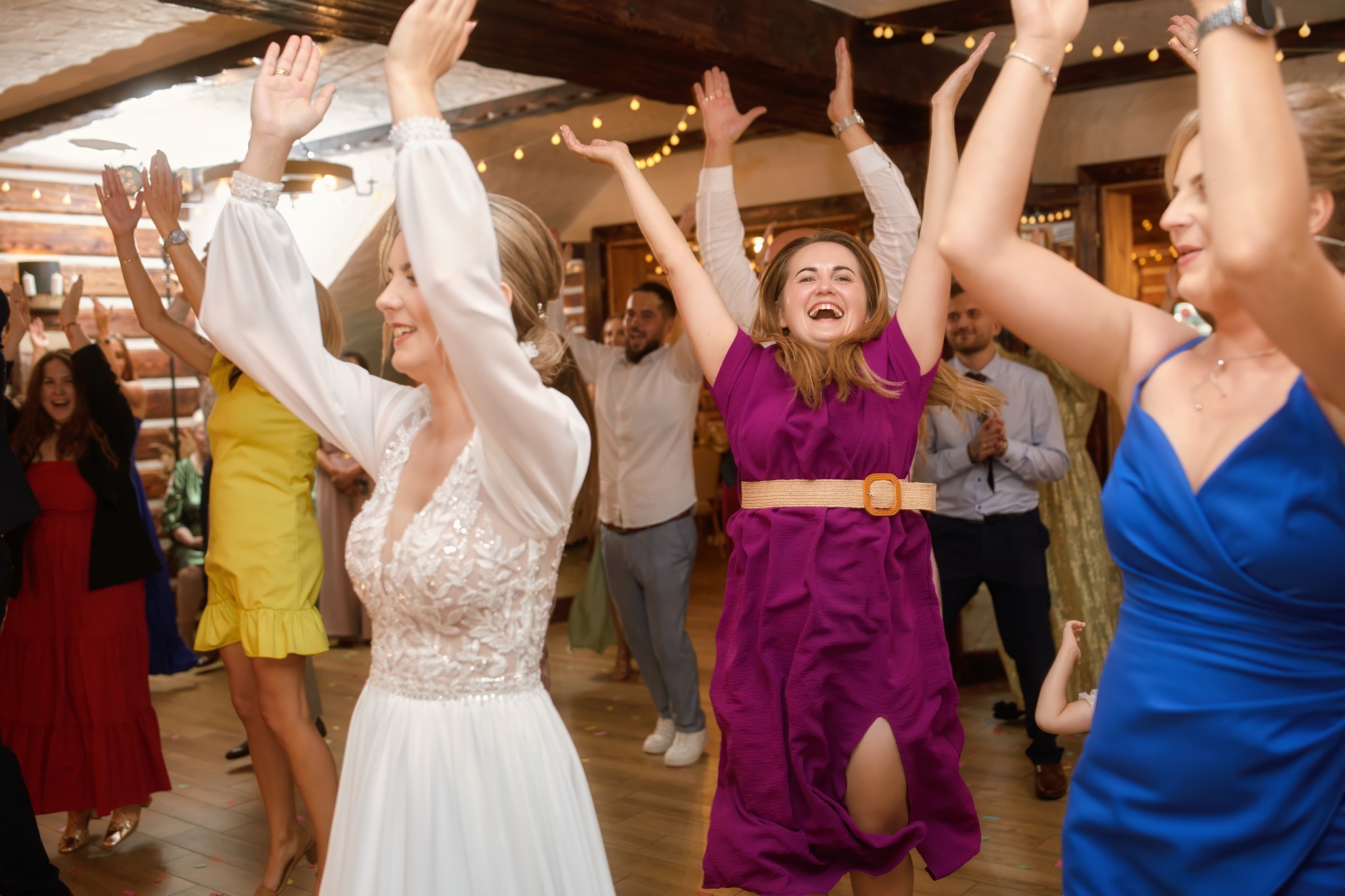 Rozbawieni goście weselni w kolorowych sukienkach tańczą z podniesionymi rękami na parkiecie. W tle drewniane elementy dekoracyjne i ciepłe oświetlenie.