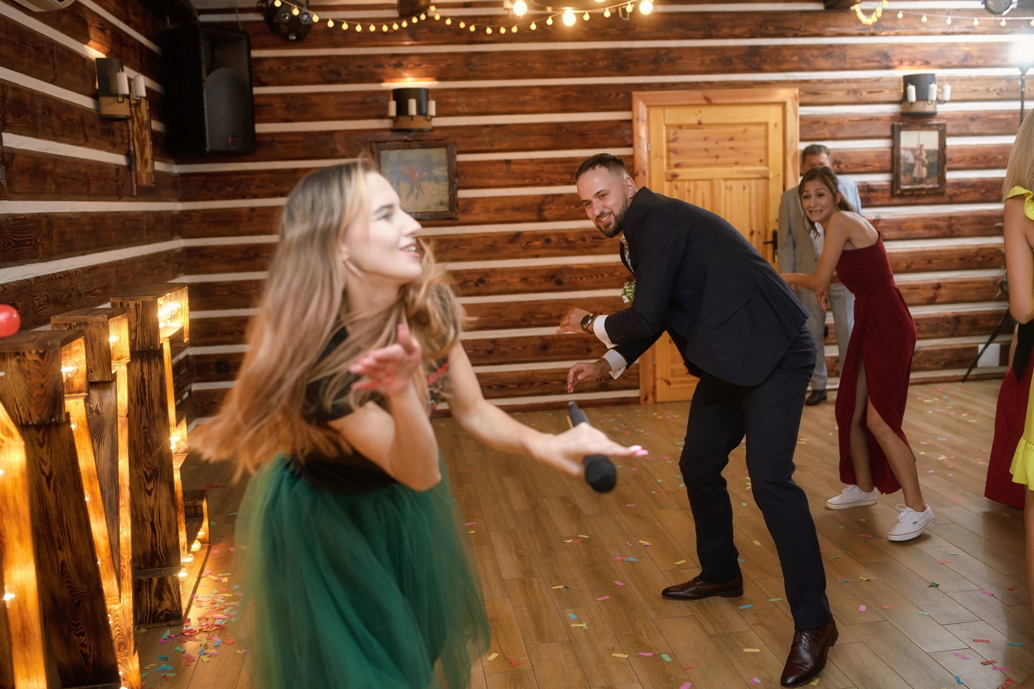 Rozbawiona para tańczy na parkiecie w rustykalnej sali balowej, z mikrofonem w dłoni. W tle goście weselni i drewniane ściany z dekoracyjnym oświetleniem.