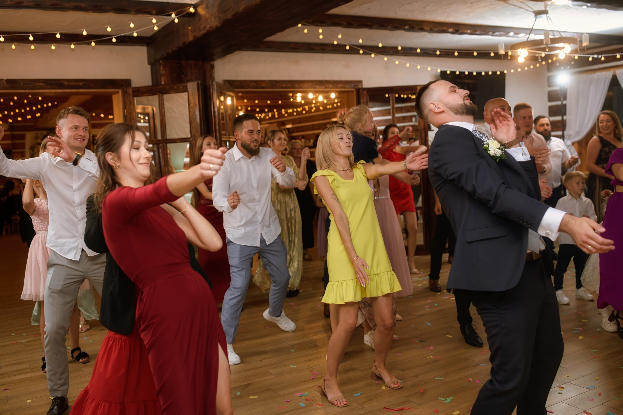 Roztańczona grupa gości weselnych w eleganckiej sali z drewnianymi elementami i girlandami świetlnymi. Wesoła atmosfera, dynamiczne ujęcie.