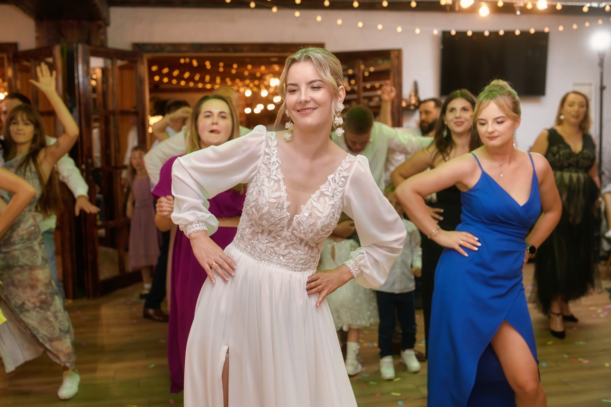 Uśmiechnięta panna młoda w białej sukni z rozcięciem tańczy z gośćmi weselnymi na parkiecie. W tle dekoracje świetlne i drewniane elementy wystroju.