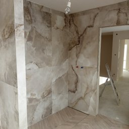 DV Solid - Wykończona łazienka z beżowymi płytkami imitującymi marmur na ścianach i podłodze ułożonej w jodełkę. Widoczne przyłącza wodne i drabina w przejściu.