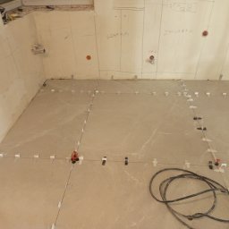 DV Solid - Układanie beżowych płytek podłogowych z użyciem systemu poziomowania. Na ścianie widoczne oznaczenia 'szafka' i 'zmywarka'.