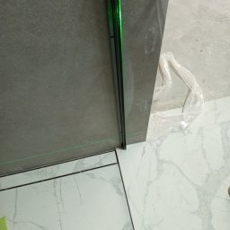 DV Solid - Układanie płytek podłogowych i ściennych z użyciem poziomicy laserowej. Widoczne płytki imitujące marmur i szare płytki ścienne. Prace wykończeniowe w toku.