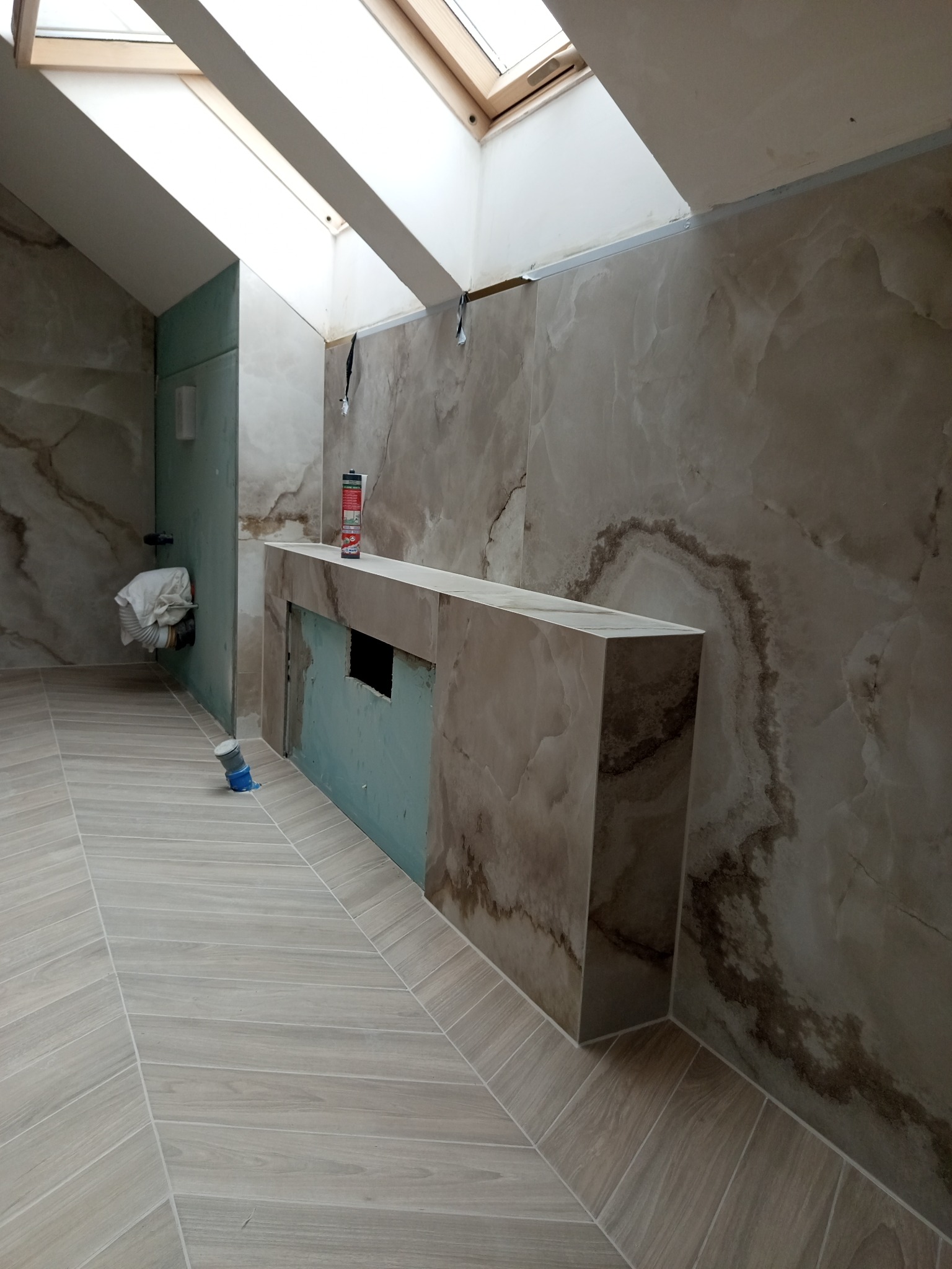 Łazienka w trakcie wykończenia: marmurowe płytki na ścianach i podłodze ułożone w jodełkę, zabudowa umywalki z widocznym otworem, okno dachowe. Etap prac wykończeniowych.
