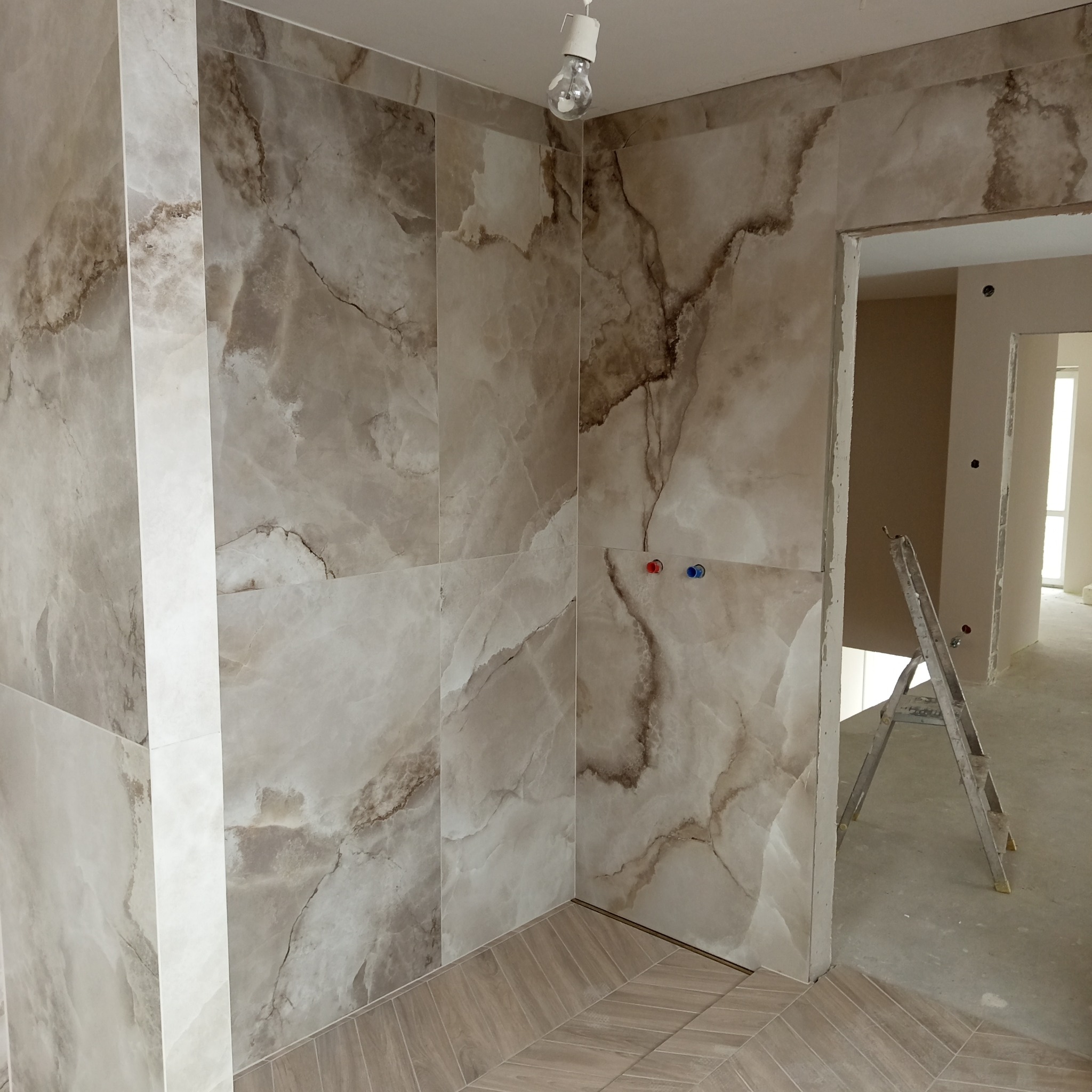 Wykończona łazienka z beżowymi płytkami imitującymi marmur na ścianach i podłodze ułożonej w jodełkę. Widoczne przyłącza wodne i drabina w przejściu.