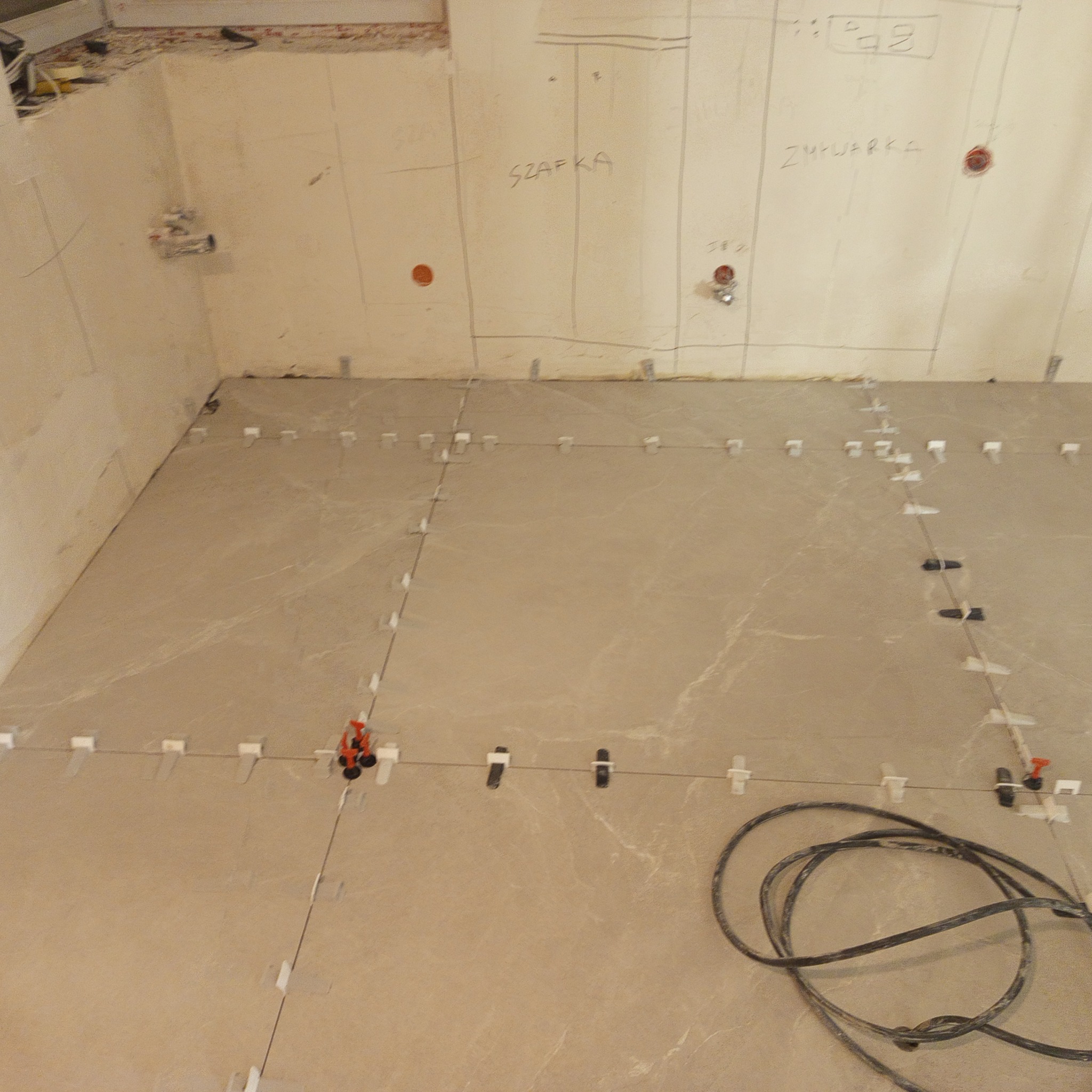 Układanie beżowych płytek podłogowych z użyciem systemu poziomowania. Na ścianie widoczne oznaczenia 'szafka' i 'zmywarka'.