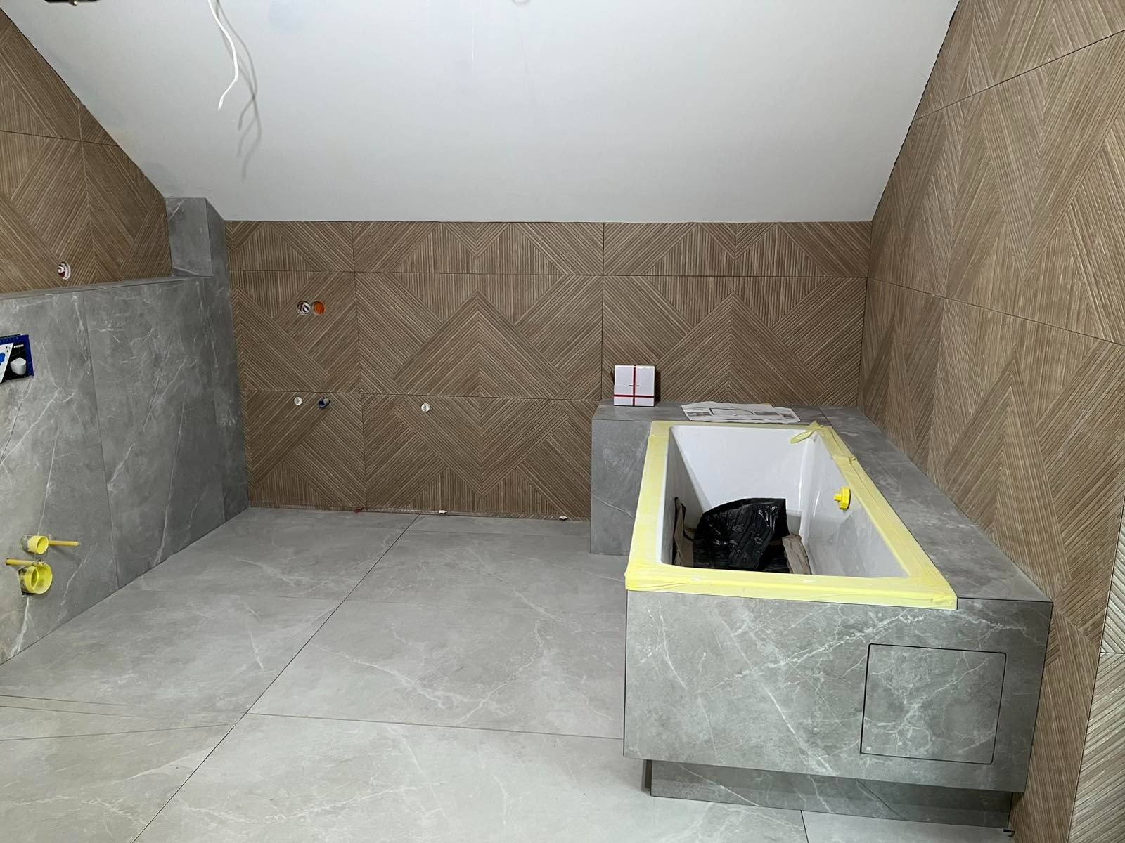 Łazienka w trakcie wykończenia: szare płytki podłogowe i ścienne imitujące drewno ułożone we wzór. Wanna obudowana płytkami, widoczne instalacje. Skos sufitu.