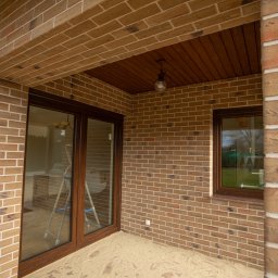 Rem-Bud Grzegorz Bernatek - Wykończony taras z ceglaną elewacją, brązowe okna i drzwi balkonowe. Widoczna drabina wewnątrz. Podłoga pokryta piaskiem. Drewniana podbitka dachowa.