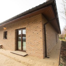 Rem-Bud Grzegorz Bernatek - Nowoczesny, parterowy dom z cegły klinkierowej w odcieniach beżu i brązu. Widoczne okna z brązowymi ramami, brązowe rynny i oświetlenie zewnętrzne. Otoczenie w stanie surowym.