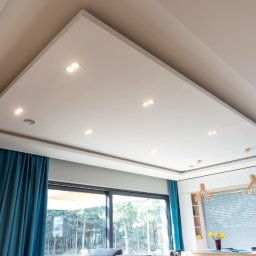 Rem-Bud Grzegorz Bernatek - Podwieszany sufit z oświetleniem punktowym w nowoczesnym salonie. Widoczne okno z turkusowymi zasłonami i fragment jadalni. Minimalistyczny design.