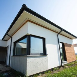 Rem-Bud Grzegorz Bernatek - Nowoczesny dom jednorodzinny z białymi ścianami i drewnianą elewacją, widok z zewnątrz, błękitne niebo w tle. Minimalistyczny design, duże okna.