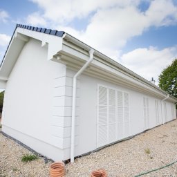 Rem-Bud Grzegorz Bernatek - Boczny widok otynkowanego na biało domu z ciemnym dachem i białymi okiennicami, otoczonego żwirowym podłożem. Widoczne rynny i fragment ogrodu.