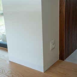 Rem-Bud Grzegorz Bernatek - Biały narożnik ściany z gładzią, listwa przypodłogowa, gniazdko elektryczne i drewniana podłoga. Fragment drewnianej ścianki działowej w tle.