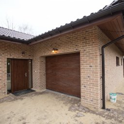Rem-Bud Grzegorz Bernatek - Nowoczesny budynek z cegły klinkierowej z brązową bramą garażową i drzwiami wejściowymi. Widoczny fragment dachu, rynny i otoczenie budynku.