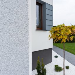 Rem-Bud Grzegorz Bernatek - Szara, ziarnista elewacja domu jednorodzinnego z oknem i ozdobnym drzewkiem na tle trawnika. Nowoczesne wykończenie zewnętrzne budynku.