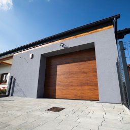 Rem-Bud Grzegorz Bernatek - Gładka, szara elewacja garażu z drewnianą bramą segmentową, oświetlona górnym światłem. Widoczny fragment kostki brukowej i ogrodzenia panelowego.