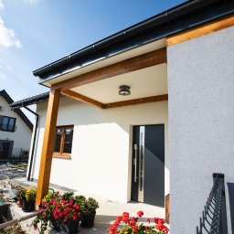 Rem-Bud Grzegorz Bernatek - Nowoczesny dom jednorodzinny z jasną elewacją i drewnianą konstrukcją werandy. Widoczna struktura tynku na ścianie i ciemne okna. W tle drugi budynek.