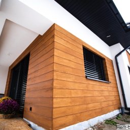 Rem-Bud Grzegorz Bernatek - Wykończona elewacja domu z drewnopodobną okładziną, czarnymi żaluzjami w oknach i białymi elementami architektonicznymi. W tle widoczny fragment ogrodu i sąsiednie budynki.