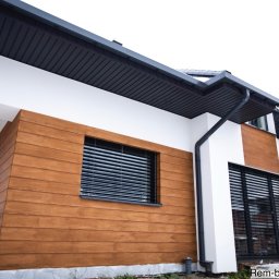 Rem-Bud Grzegorz Bernatek - Nowoczesny dom z elewacją w odcieniach bieli i drewna, z czarnymi akcentami dachu i okien. Widoczne detale wykończenia, takie jak rynny i żaluzje okienne.