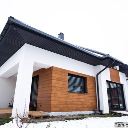 Rem-Bud Grzegorz Bernatek - Nowoczesny dom jednorodzinny z drewnianą elewacją i białymi ścianami, widziany z niskiej perspektywy w zimowej scenerii. Widoczne czarne okna i dach.