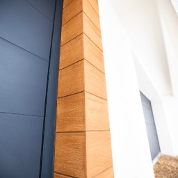 Rem-Bud Grzegorz Bernatek - Eleganckie wykończenie elewacji z drewnopodobnych paneli przy drzwiach wejściowych. Detal narożnika z widoczną strukturą drewna i precyzyjnym łączeniem.