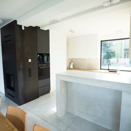 Rem-Bud Grzegorz Bernatek - Nowoczesna kuchnia z wyspą w odcieniach bieli, szarości i czerni. Minimalistyczne szafki, duże okno z widokiem na zieleń. Elementy wykończenia imitujące beton.