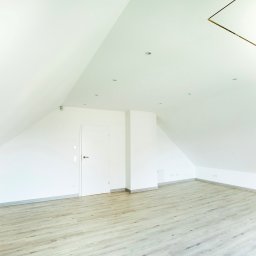 Rem-Bud Grzegorz Bernatek - Jasne, świeżo wykończone poddasze z białymi ścianami i panelami podłogowymi w odcieniach szarości. Widoczne skosy, białe drzwi i oświetlenie punktowe.